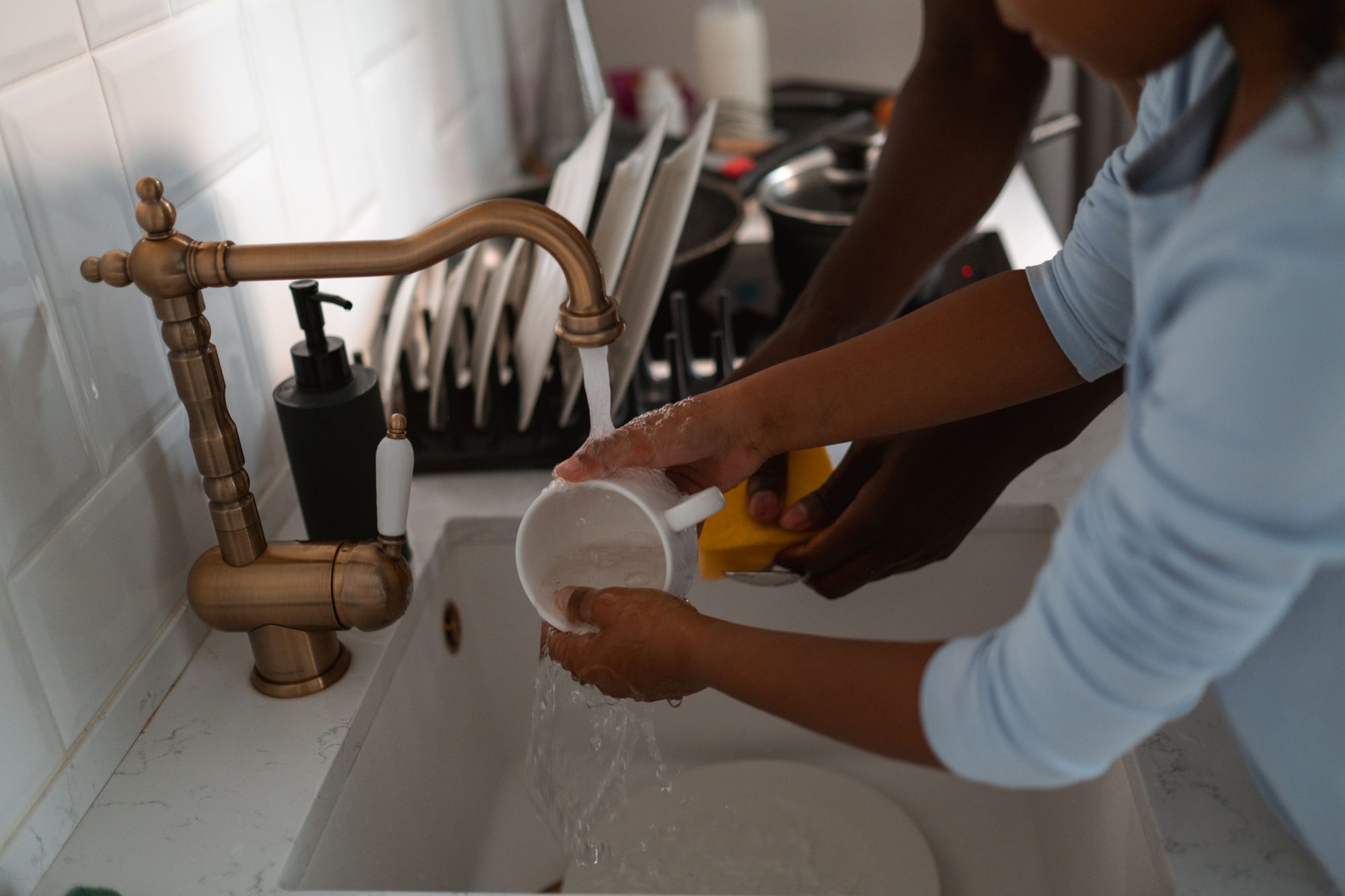 Hands rinsing a cup under an antique brass swan-neck kitchen tap over a white ceramic sink